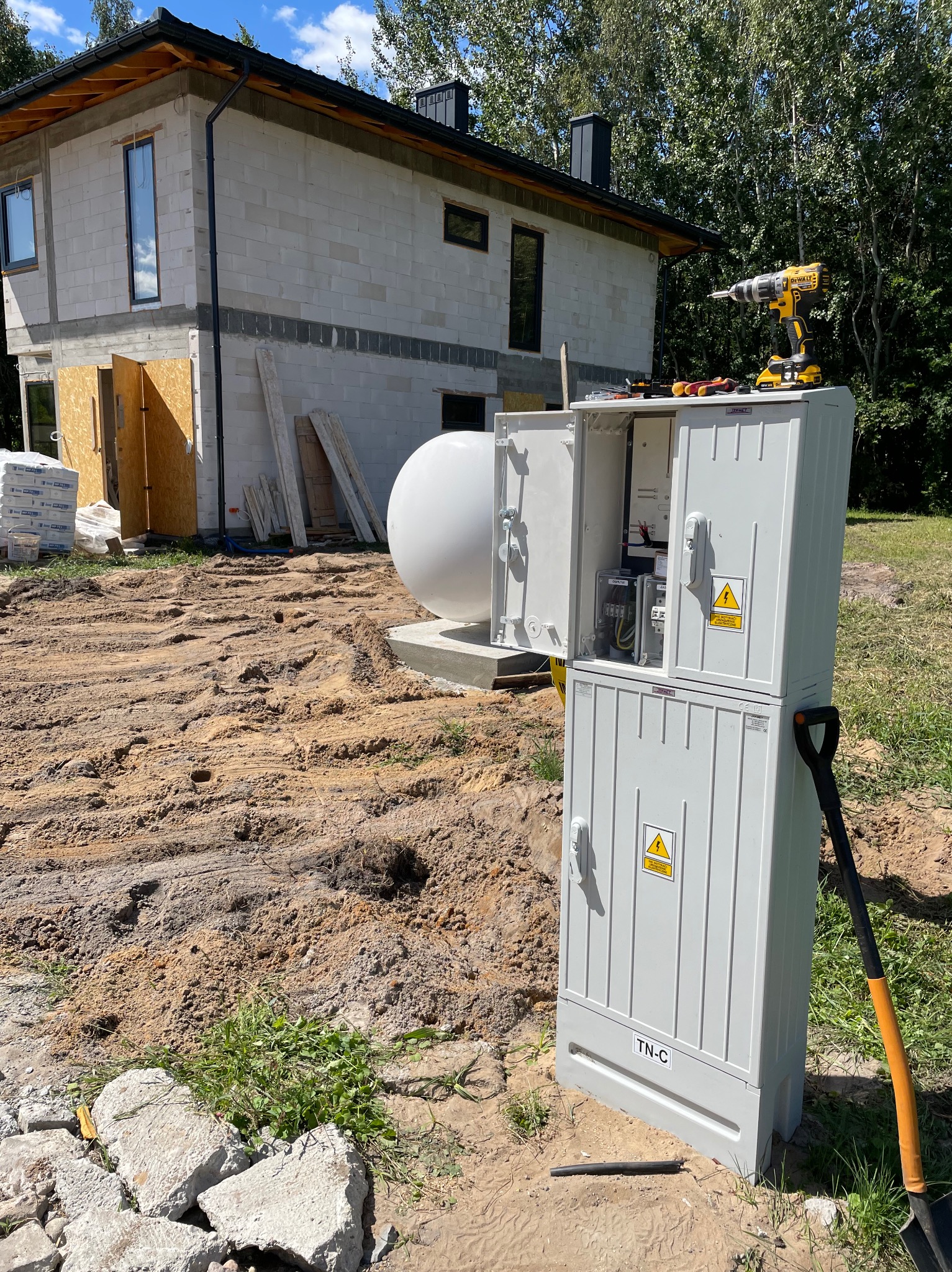 Dwuczęściowa, otwarta skrzynka elektryczna TN-C z ostrzeżeniem o napięciu, stojąca na piaszczystym gruncie budowlanym przed domem w stanie surowym, z widocznym zbiornikiem gazu, w tle drzewa...