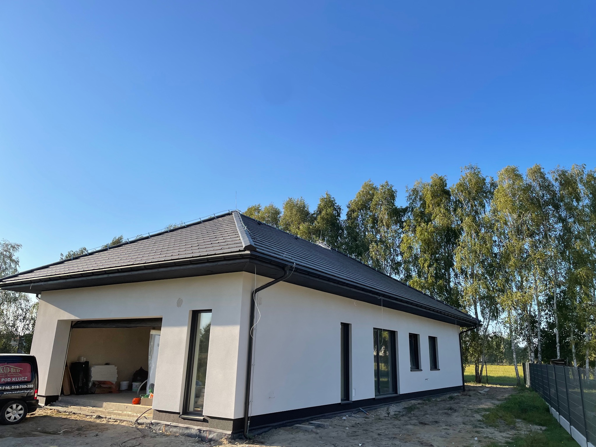 Nowoczesny, parterowy dom z szarą dachówką i białą elewacją, widoczna instalacja odgromowa na dachu, błękitne niebo w tle, drzewa liściaste wzdłuż ogrodzenia.