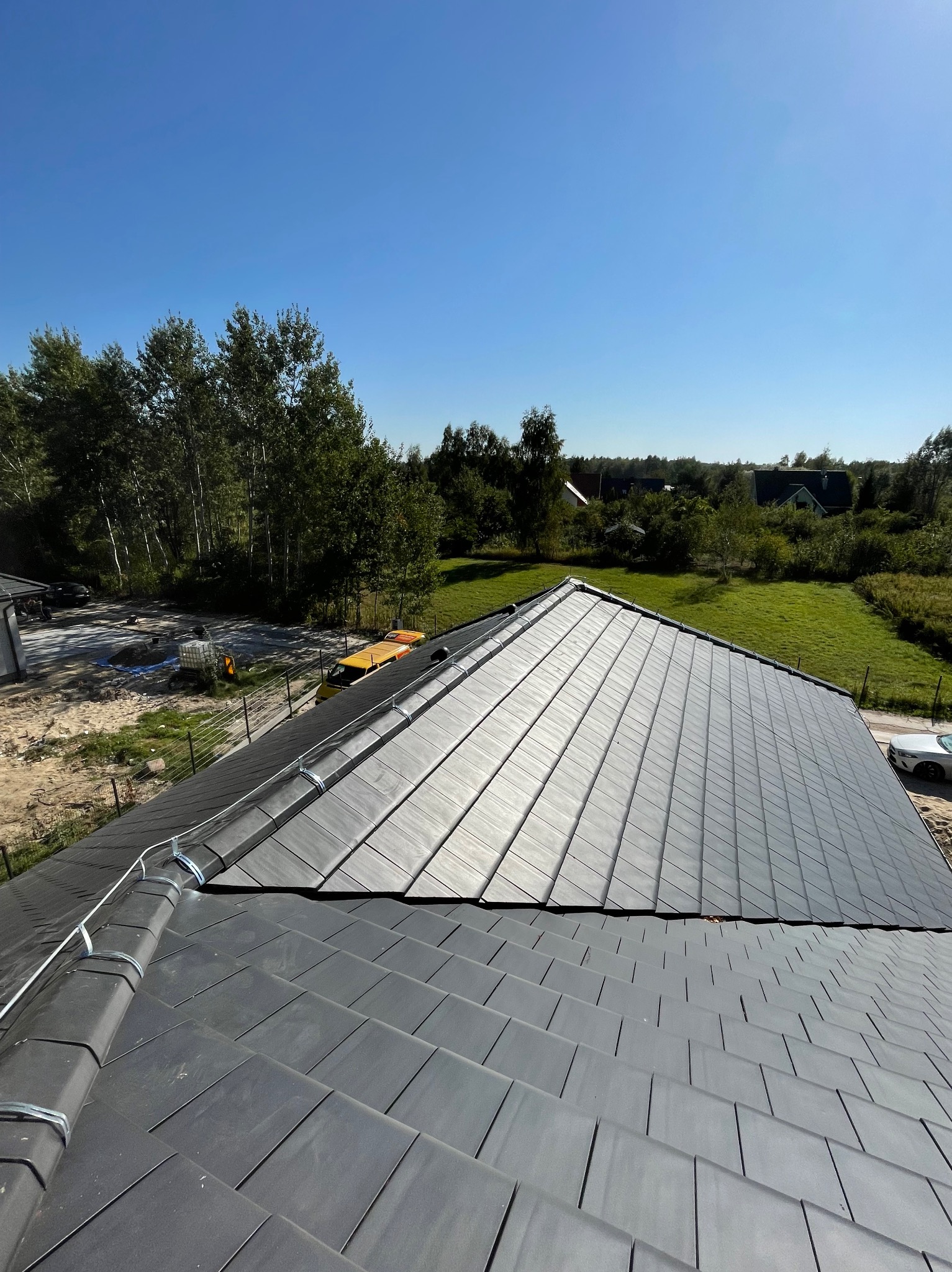 Widok z góry na dach pokryty szarą dachówką z zainstalowaną instalacją odgromową, widoczne metalowe uchwyty i przewody, w tle zieleń i błękitne niebo.