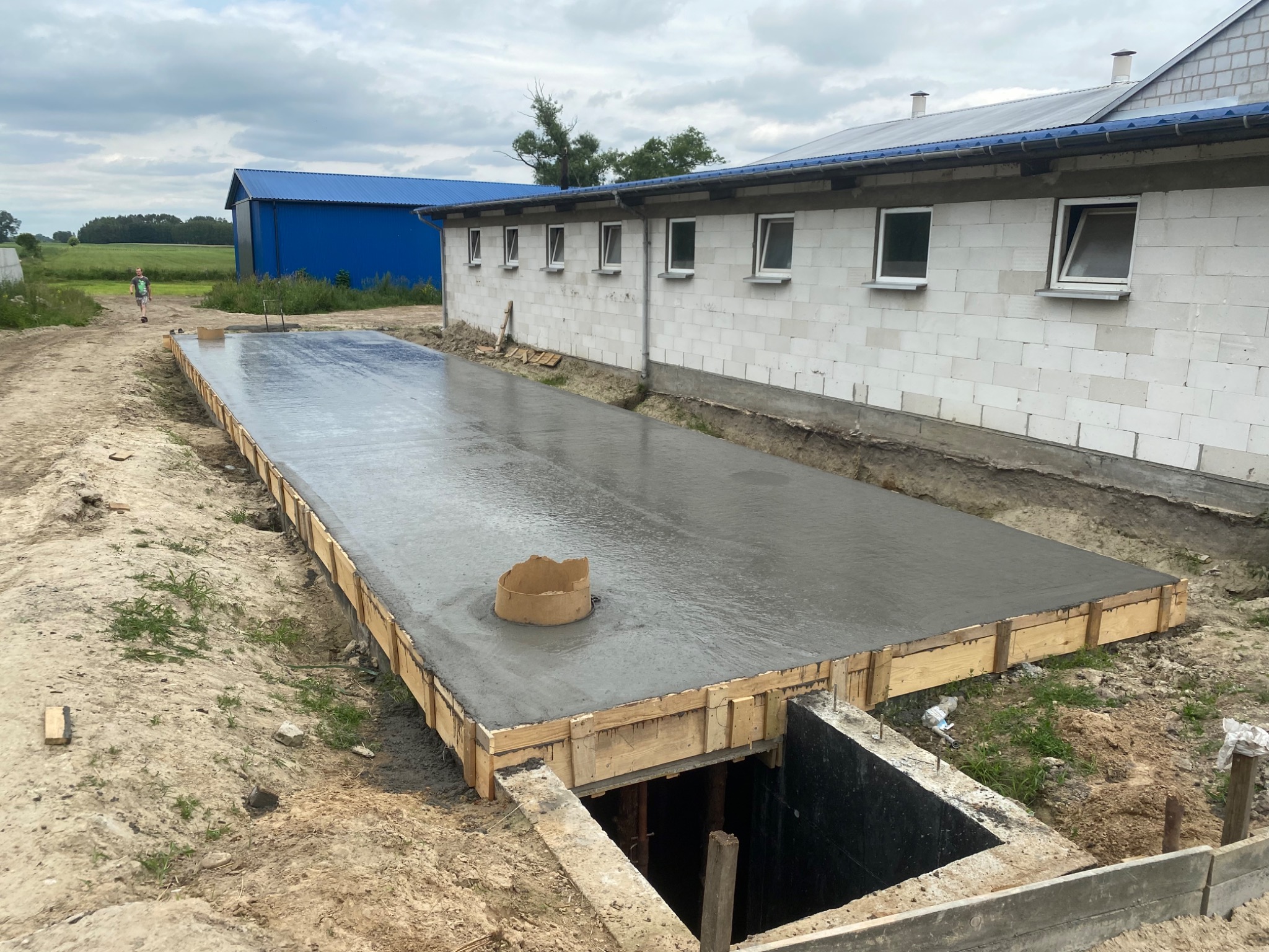 Wylany fundament pod budynek gospodarczy z widoczną studzienką rewizyjną i szalunkiem drewnianym, w tle budynek z białego bloczka i niebieska hala.