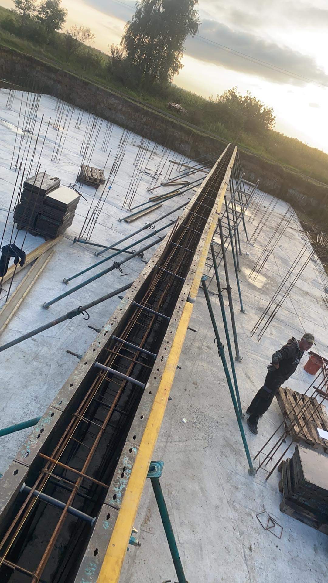 Widok z góry na plac budowy z przygotowaną formą i zbrojeniem pod wylewkę fundamentową, widoczne podpory i pracownik w oddali.