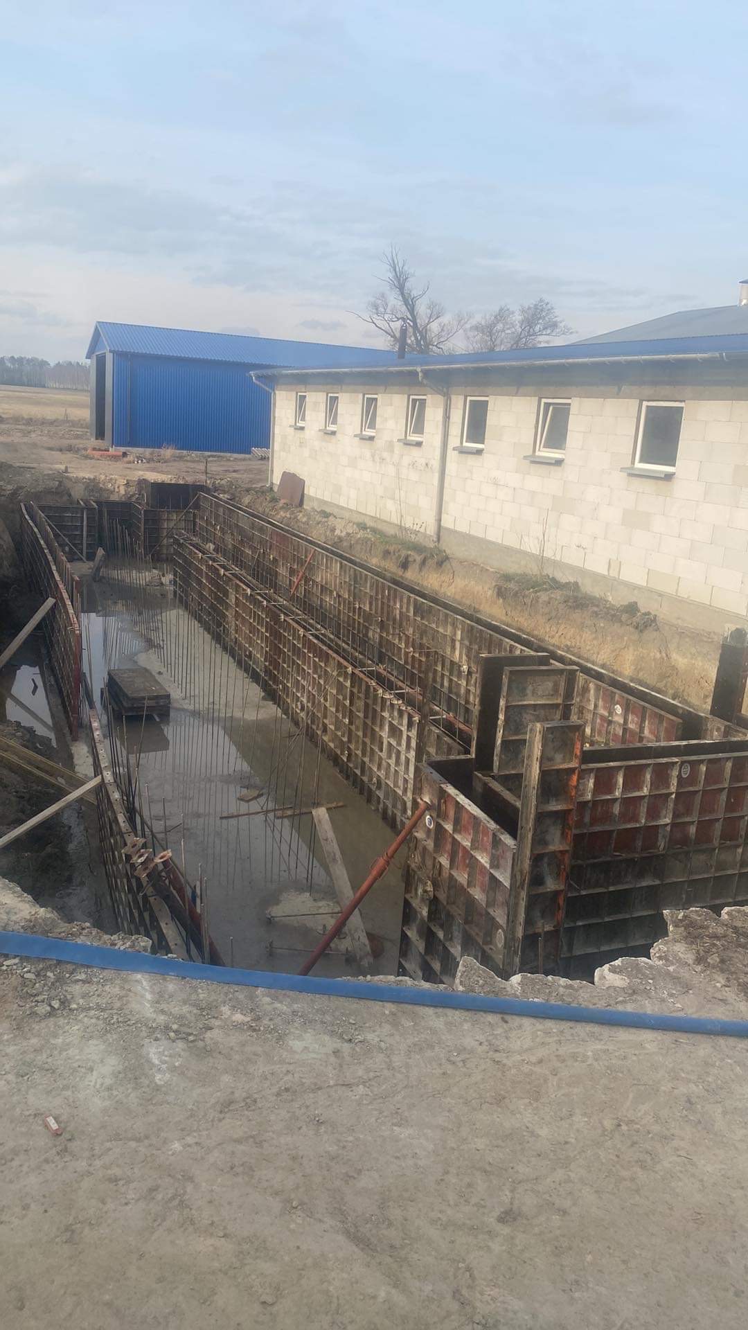 Wykonanie szalunków pod fundamenty budynku gospodarczego, widoczne stalowe pręty zbrojeniowe i woda w wykopie, w tle murowany budynek i hala z blachy trapezowej.