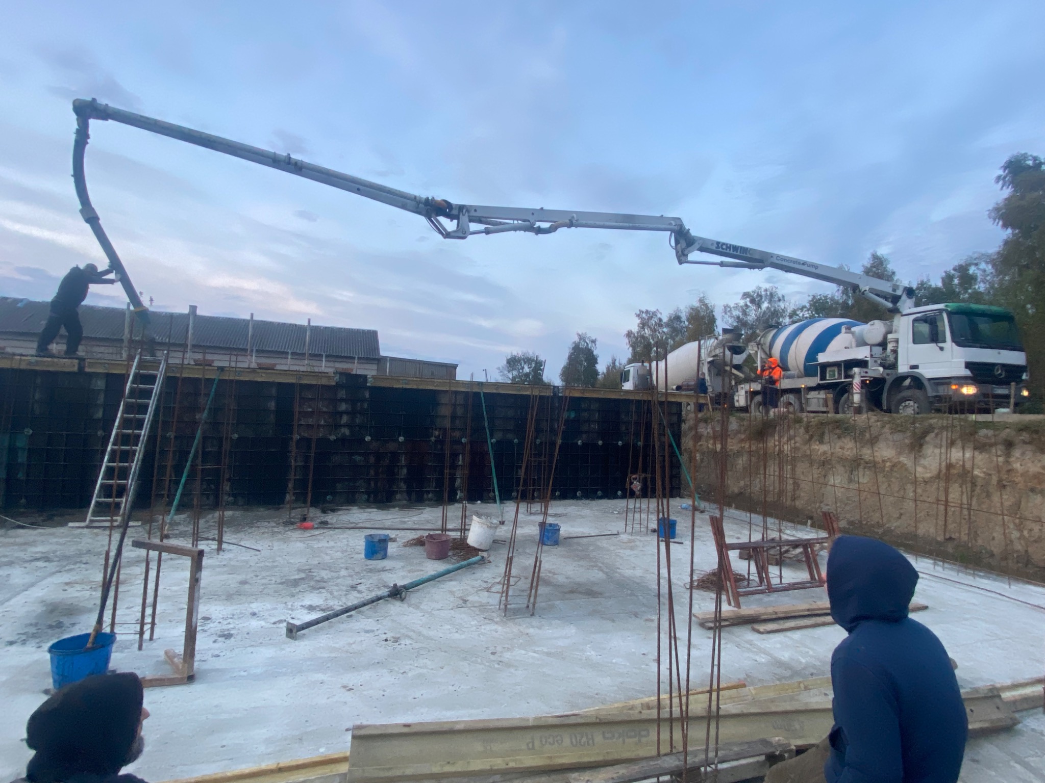 Wylewanie betonu na fundament płyty budynku za pomocą pompy do betonu, widoczne zbrojenie i szalunki, w tle betonomieszarka oraz pracownicy.