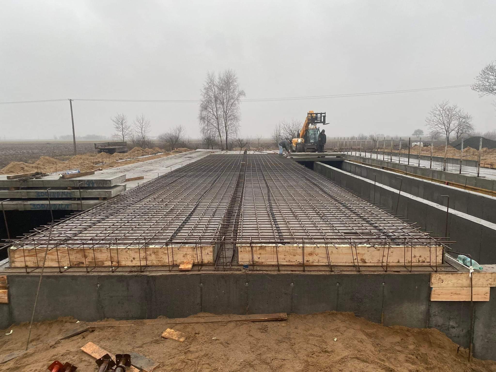 Zbrojenie płyty fundamentowej z użyciem drewnianego szalunku, widoczny pracownik i podnośnik teleskopowy w tle, pochmurny dzień.