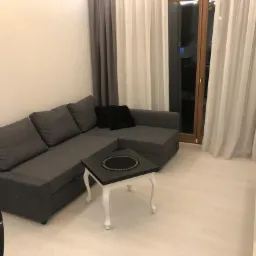 Szary narożnik z poduszkami i mały czarny stolik na białych nóżkach w salonie z zasłonami, obok stoi odkurzacz pionowy.