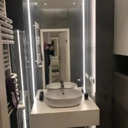 Nowoczesna łazienka z podświetlanym lustrem, białą umywalką nablatową i szarą płytką na ścianie. W lustrze odbija się osoba robiąca zdjęcie.