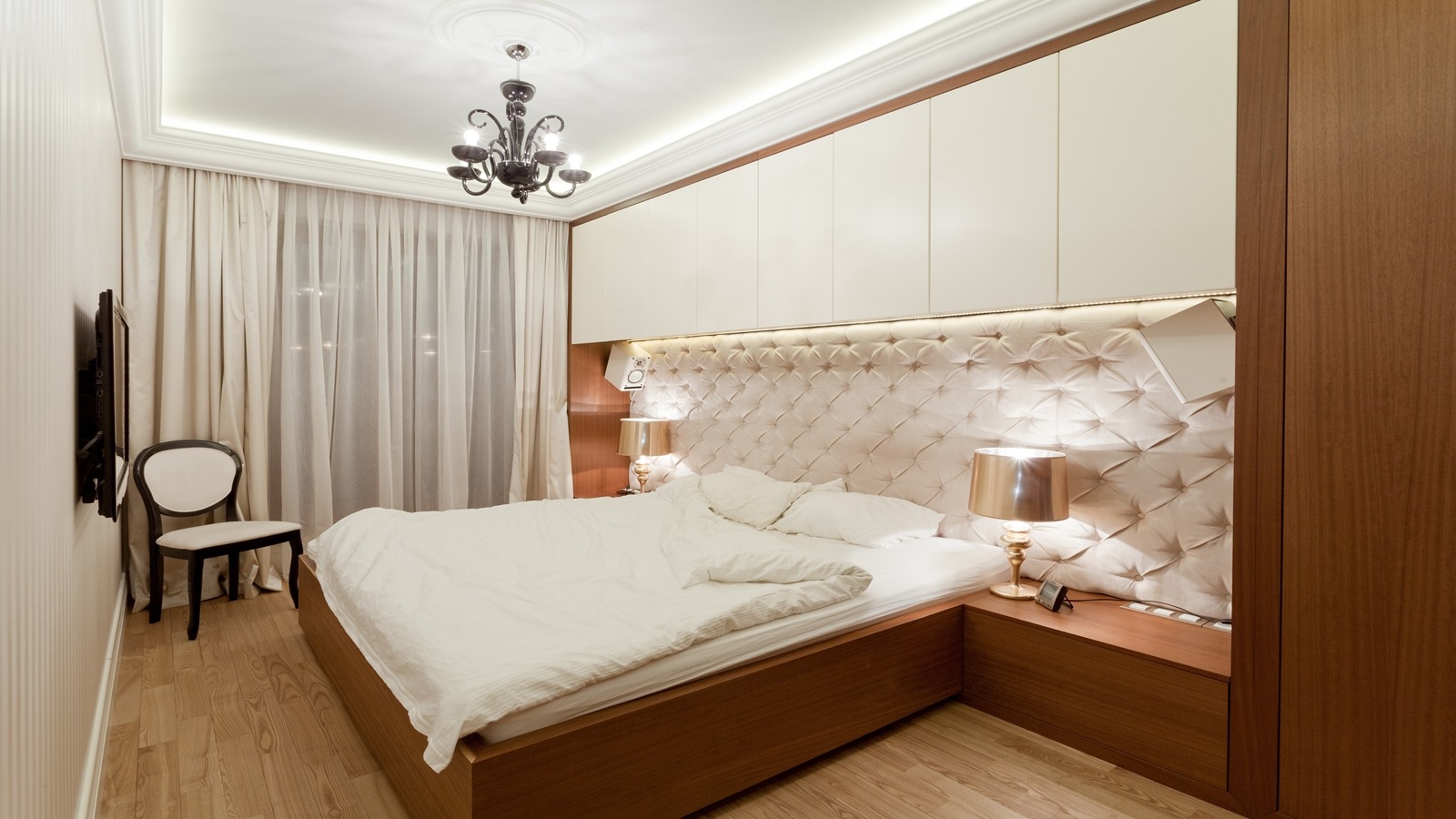 Elegancka sypialnia z łóżkiem z tapicerowanym zagłówkiem w romby, oświetleniem LED i szafkami wiszącymi, uzupełniona o klasyczny żyrandol i krzesło w stylu retro.