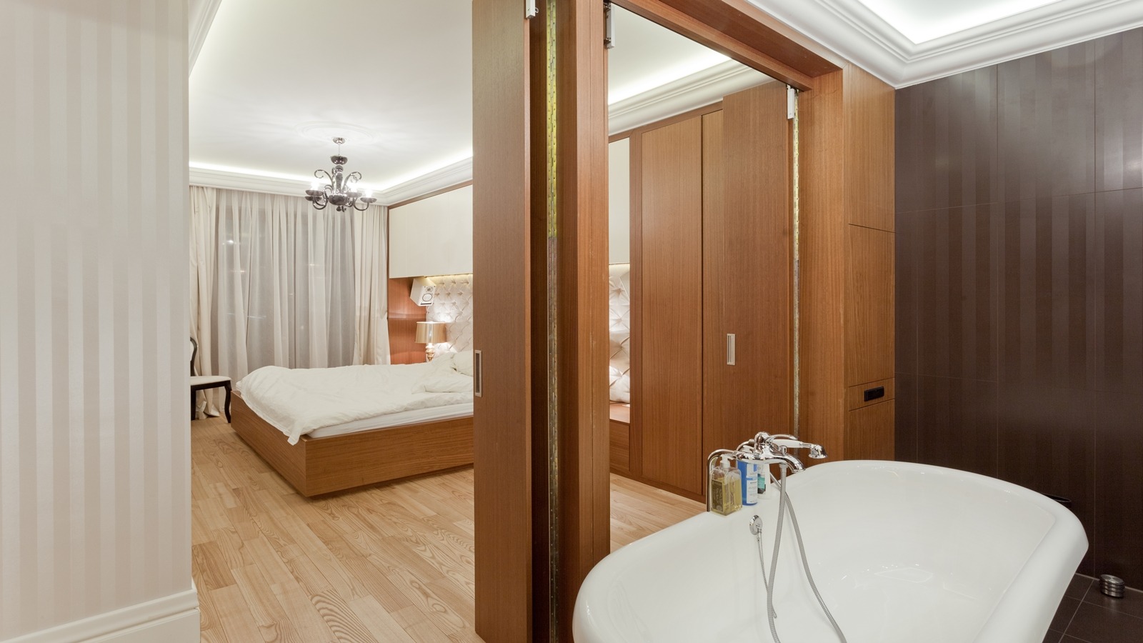 Widok na sypialnię z łazienki, drewniana podłoga i szafa z przesuwnymi drzwiami, biała wanna wolnostojąca z chromowaną armaturą.