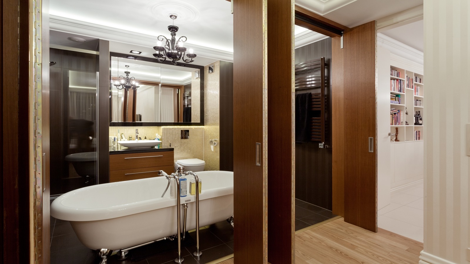 Elegancka łazienka z wolnostojącą wanną na srebrnych nóżkach, umywalką nablatową i przesuwnymi drzwiami z drewna, oświetlona dekoracyjnym żyrandolem.