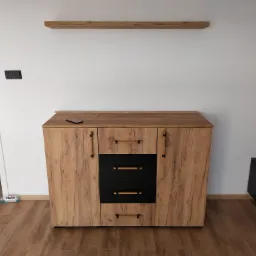 Komoda z drewnopodobnymi frontami i czarnymi szufladami, uzupełniona o wiszącą półkę w tym samym odcieniu, smartfon leżący na blacie, widoczna butelka z napojem na podłodze.