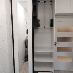 Biała szafa przesuwna z czarnymi obramowaniami, jedno skrzydło z lustrem, widoczne wnętrze z półkami, drążkiem i wysuwaną szufladą; w lustrze odbicie pokoju z sofą i wzorzystą tapetą.