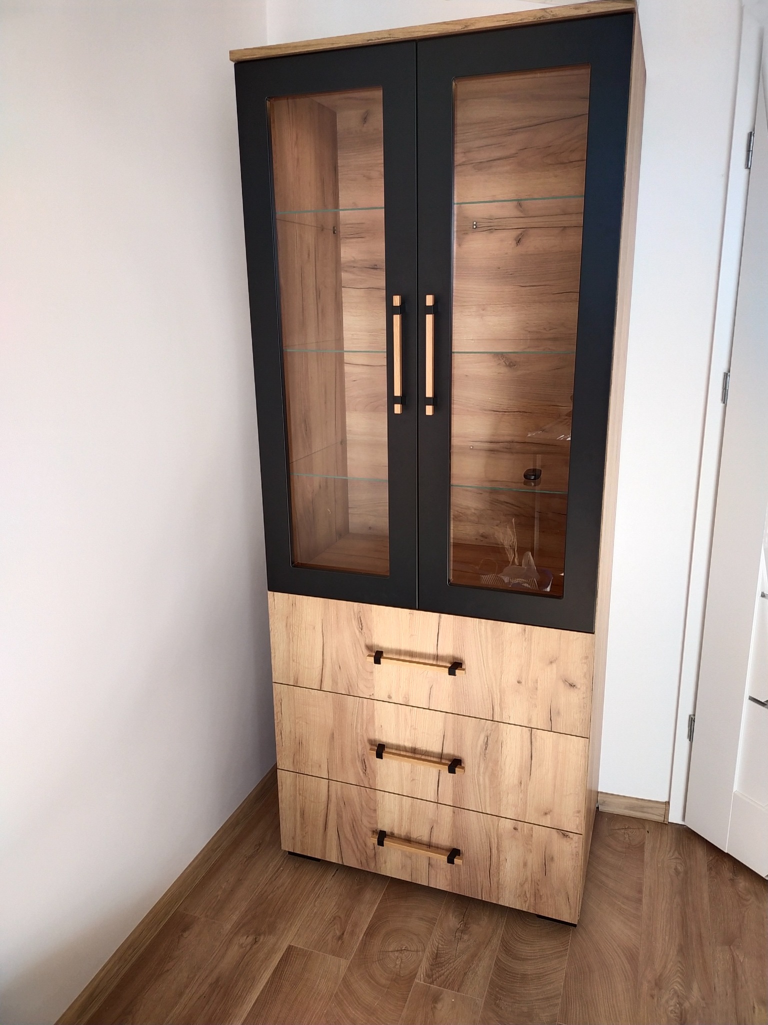 Wysoka witryna z dwoma przeszklonymi drzwiami w czarnej ramie i trzema szufladami z drewnopodobnym dekorem, stojąca na panelach podłogowych w rogu pokoju.