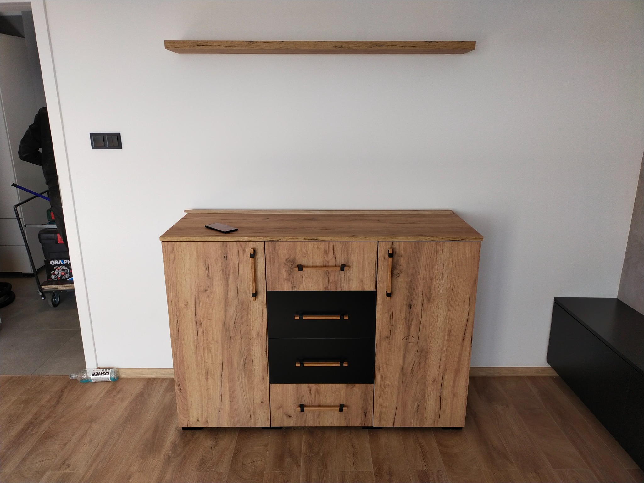 Komoda z drewnopodobnymi frontami i czarnymi szufladami, uzupełniona o wiszącą półkę w tym samym odcieniu, smartfon leżący na blacie, widoczna butelka z napojem na podłodze.