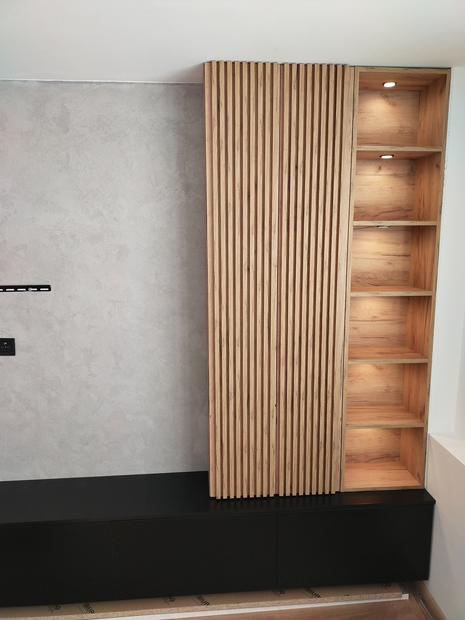 Nowoczesna zabudowa ściany z czarną szafką RTV, drewnianymi lamelami i regałem z podświetlanymi półkami.