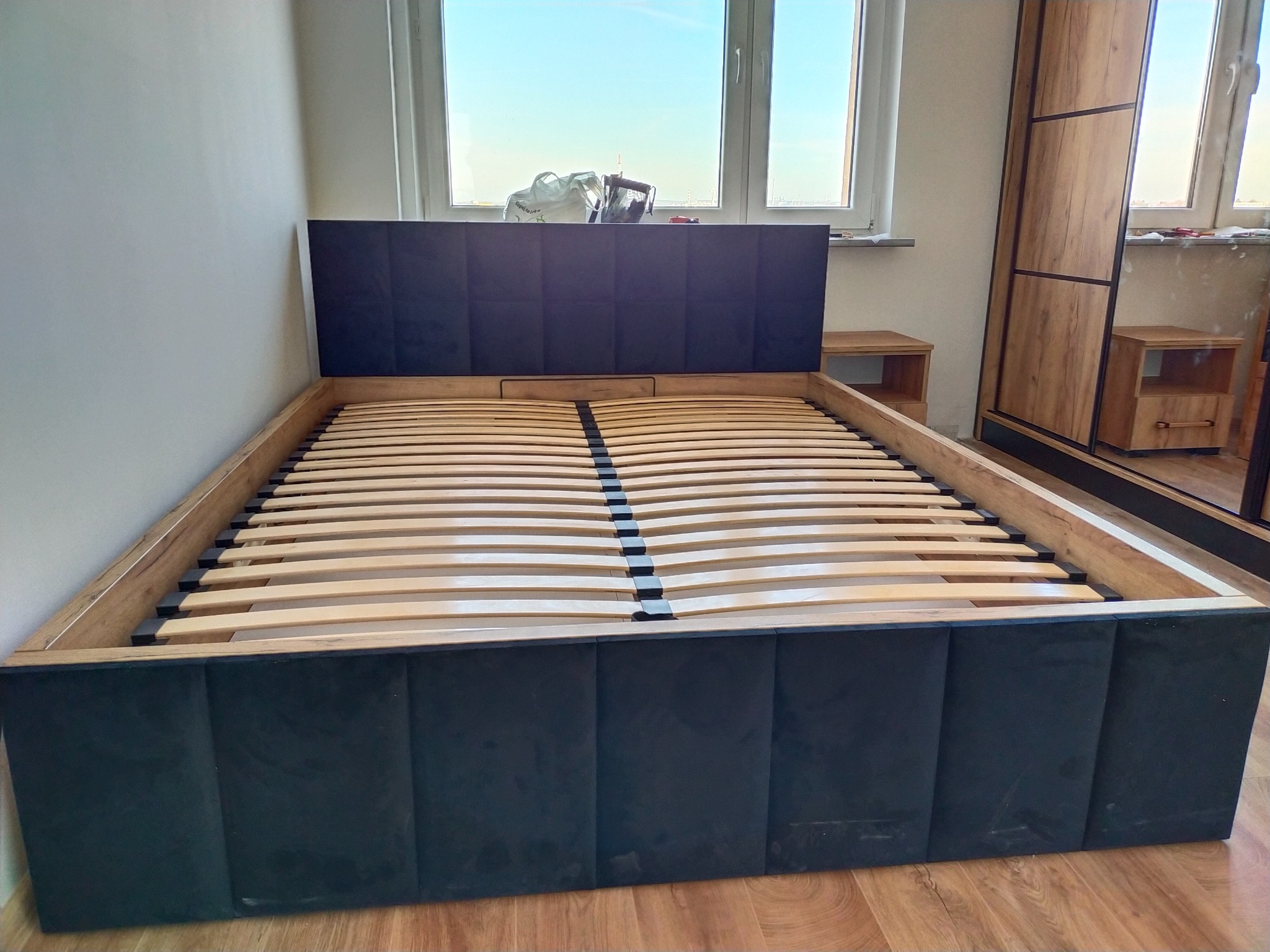 Łóżko z tapicerowanym wezgłowiem i ramą, drewniany stelaż listewkowy, obok szafka nocna i szafa przesuwna z drewnopodobnymi frontami.