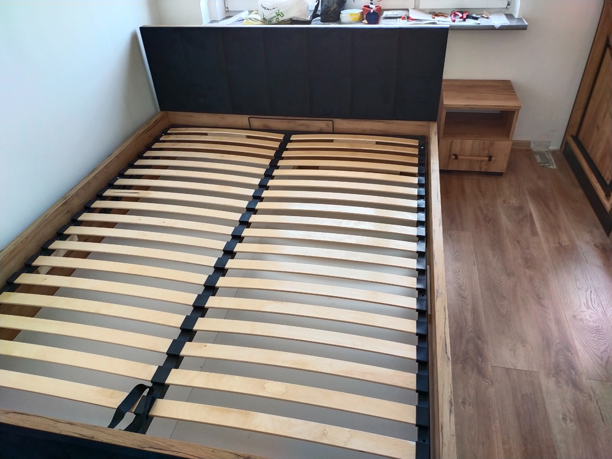 Drewniana rama łóżka z czarnym zagłówkiem i drewnianym stelażem, widoczna szafka nocna z drewna po prawej stronie, podłoga z paneli imitujących drewno.