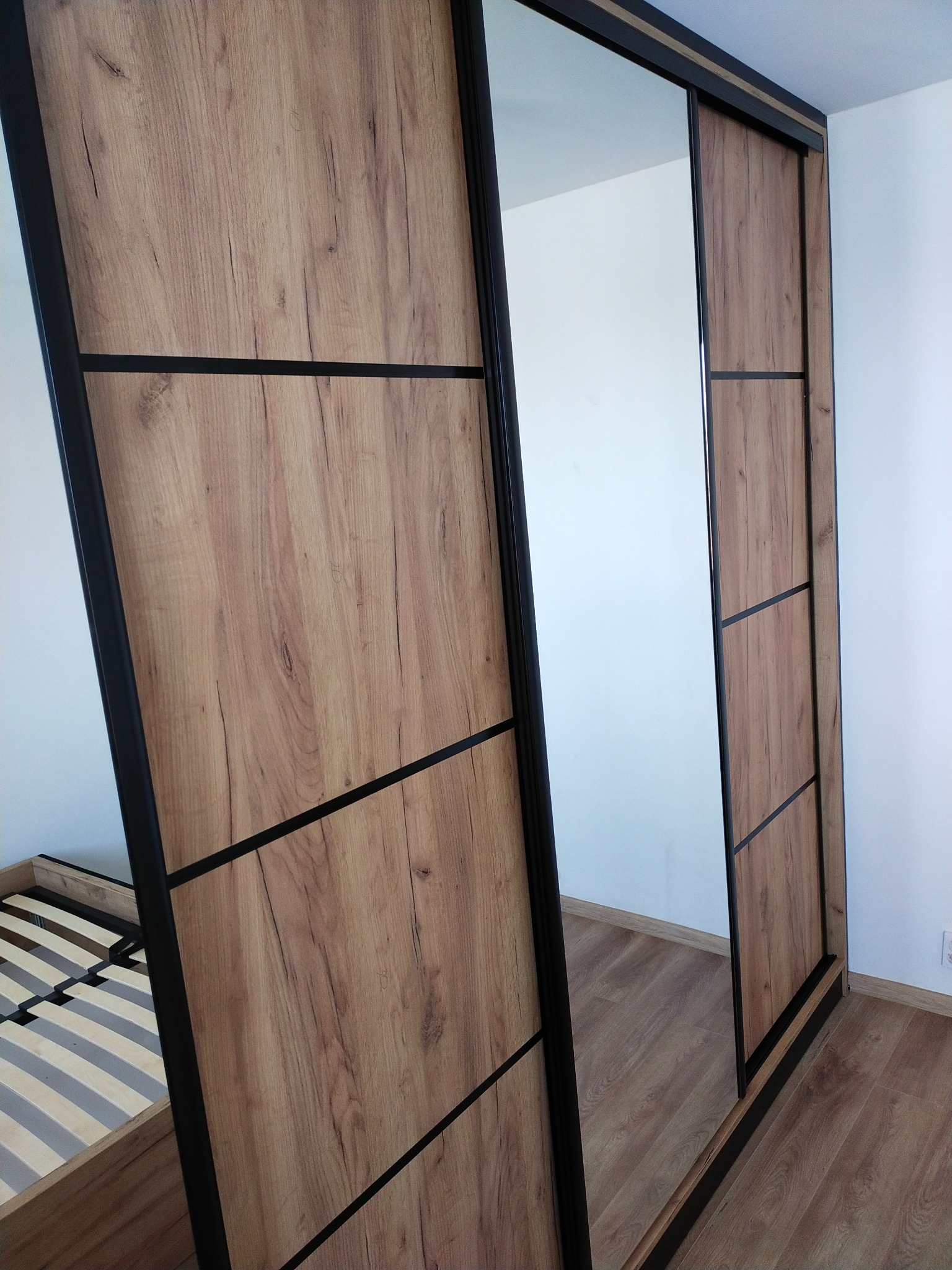 Nowoczesna szafa przesuwna z lustrem i drewnianymi panelami w odcieniach brązu, z czarnymi elementami konstrukcyjnymi, widoczna częściowo rama łóżka w tle.