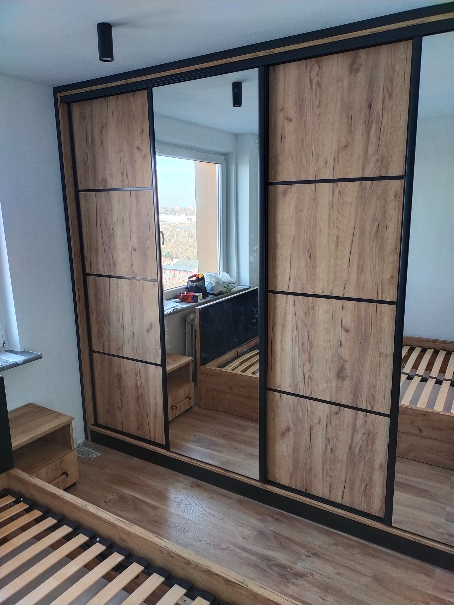 Nowoczesna szafa przesuwna z lustrzanymi drzwiami o drewnianym wzorze, czarne obramowanie, widoczne elementy łóżka i szafki nocnej w pokoju, oświetlenie sufitowe