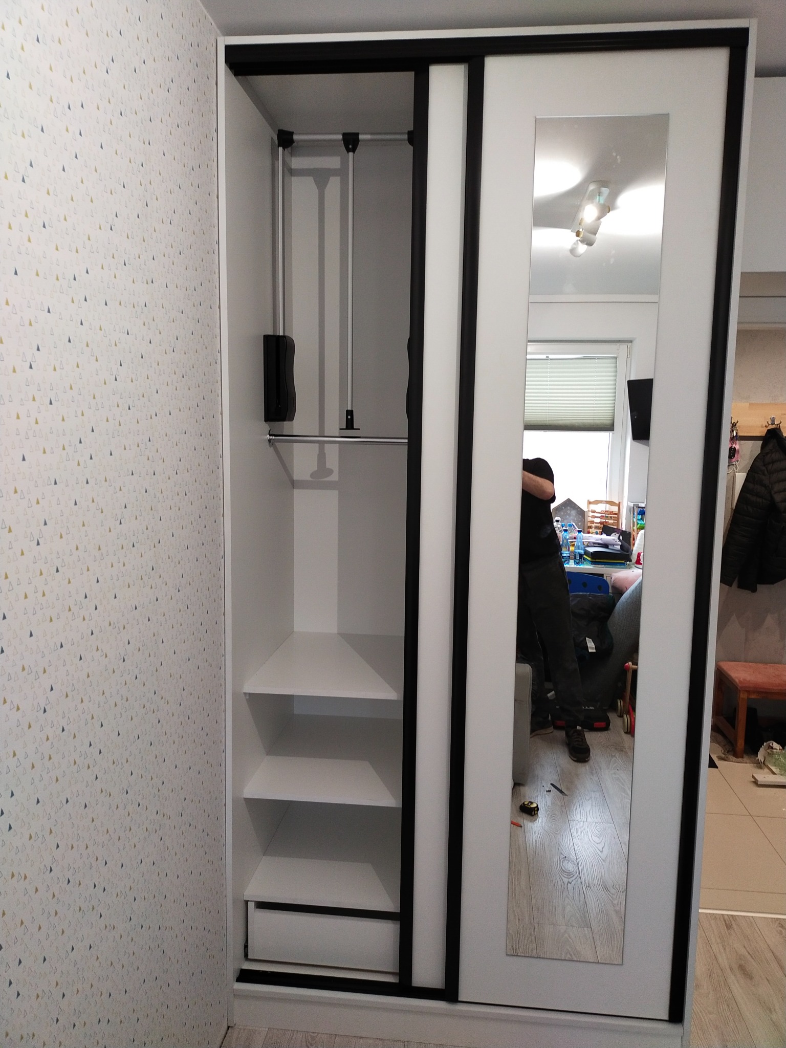 Wnętrze białej szafy z przesuwnymi drzwiami z lustrem, widoczne półki, drążek na ubrania i szuflada, odbicie osoby w lustrze.