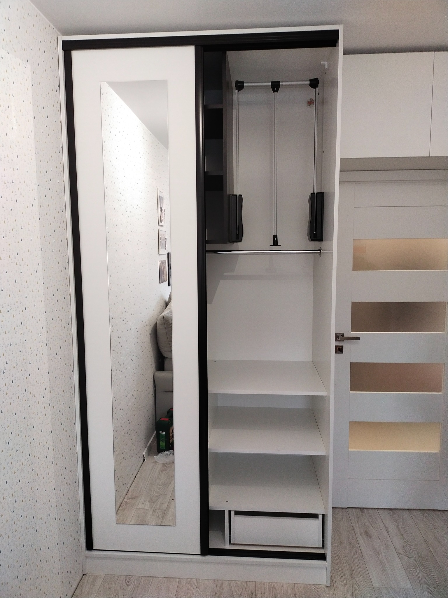 Biała szafa przesuwna z czarnymi obramowaniami, jedno skrzydło z lustrem, widoczne wnętrze z półkami, drążkiem i wysuwaną szufladą; w lustrze odbicie pokoju z sofą i wzorzystą tapetą.