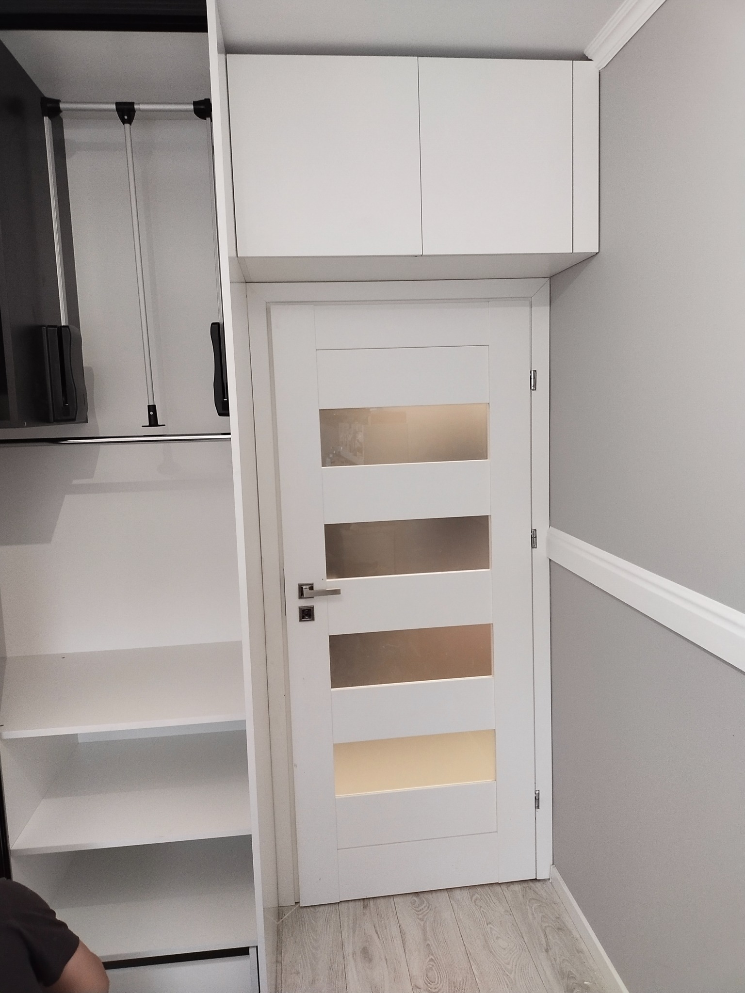 Biała szafa na wymiar z systemem drążków i półek, obok drzwi wewnętrzne z przeszkleniami i szafki wiszące nad nimi, szara ściana z białą listwą przypodłogową.