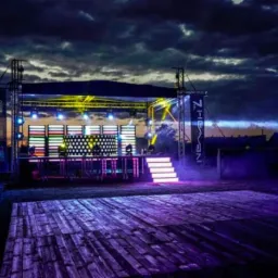 Scena plenerowa oświetlona kolorowymi światłami, z drewnianym parkietem tanecznym na pierwszym planie i zachodzącym słońcem widocznym na horyzoncie.