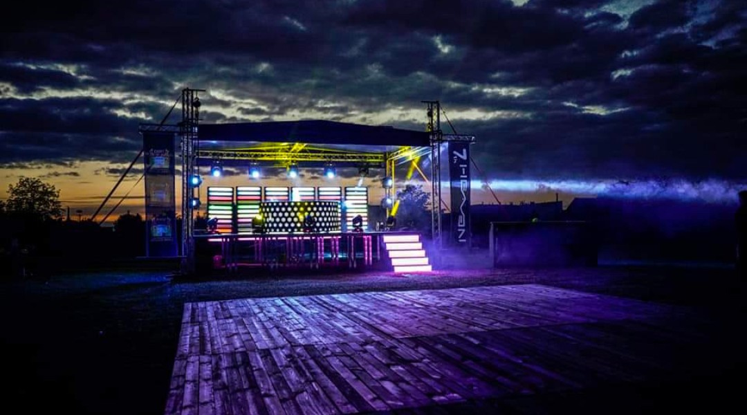 Scena plenerowa oświetlona kolorowymi światłami, z drewnianym parkietem tanecznym na pierwszym planie i zachodzącym słońcem widocznym na horyzoncie.