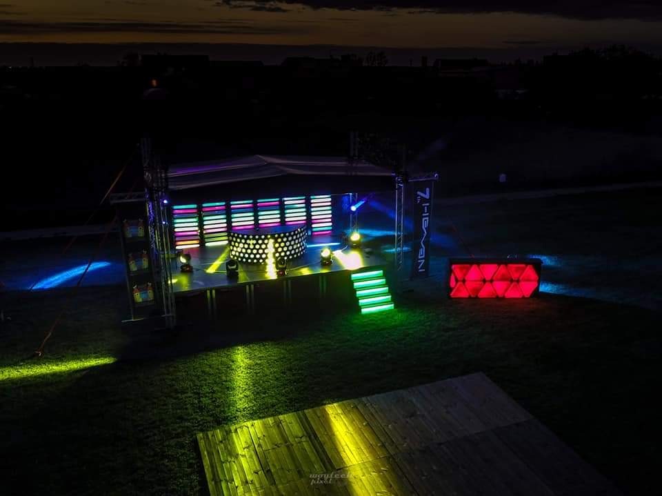 Scena eventowa z podświetleniem LED, widok z góry, oświetlenie skierowane na trawę, banner firmy po prawej stronie sceny, ciemne niebo w tle.