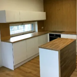 Nowoczesna kuchnia z białymi szafkami i drewnianym blatem, wyspa kuchenna z blatem z klejonego drewna, ściana wykończona panelami drewnopodobnymi.