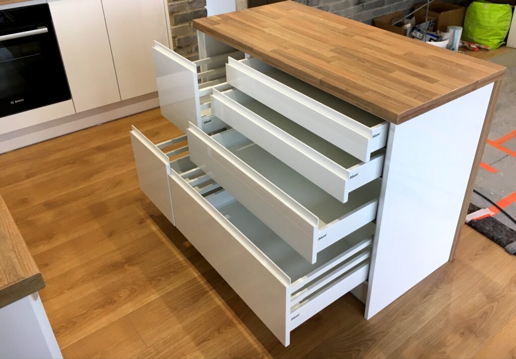 Nowoczesna wyspa kuchenna z białymi frontami i drewnianym blatem, wysuwane szuflady z systemem Blum, widoczna podłoga z paneli, w tle ściana z cegły.