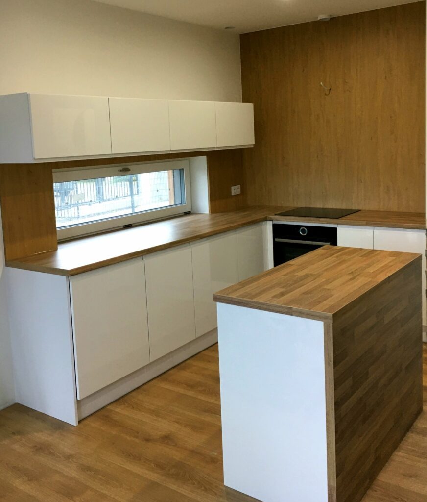 Nowoczesna kuchnia z białymi szafkami i drewnianym blatem, wyspa kuchenna z blatem z klejonego drewna, ściana wykończona panelami drewnopodobnymi.