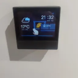 Czarny panel sterowania domowego z wyświetlaczem pokazującym temperaturę, godzinę i prognozę pogody, zamontowany na białej ścianie.