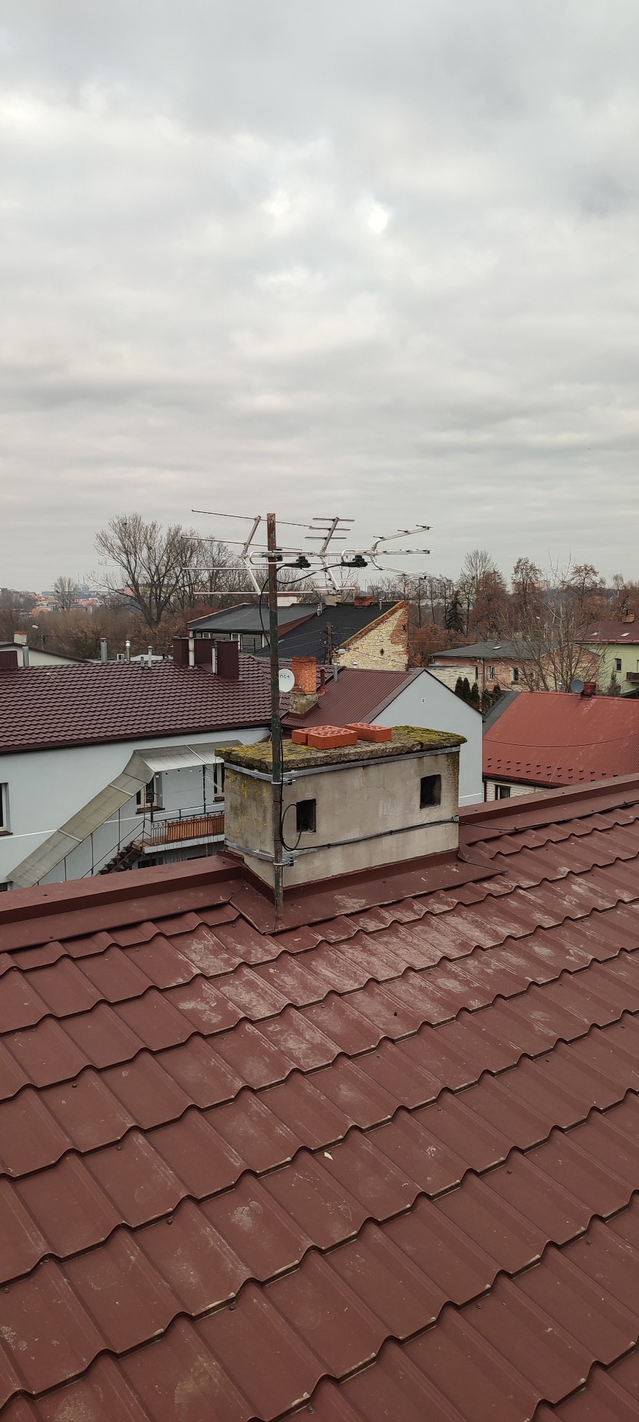 Antena Yagi-Uda zamontowana na kominie z cegłami na dachu z dachówki ceramicznej w tle zabudowa miejska pod pochmurnym niebem.