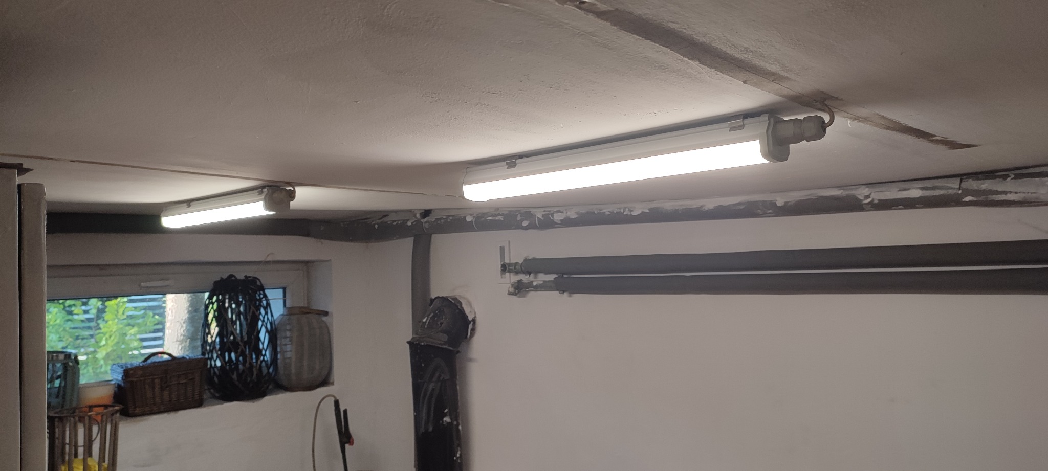 Dwie podłużne lampy fluorescencyjne zamontowane na suficie w pomieszczeniu z widocznymi rurami instalacyjnymi i oknem z dekoracjami.