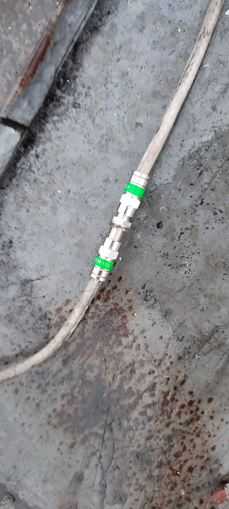 Kabel antenowy koncentryczny z zielonymi oznaczeniami 'FILTER 113' i złączkami, leżący na szarej, zardzewiałej powierzchni.