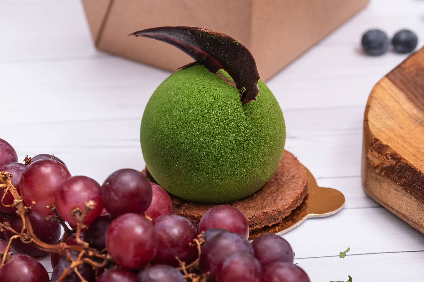 Zielony deser w kształcie kuli z czekoladową dekoracją, ułożony na czekoladowym spodzie i złotej podkładce, otoczony kiścią czerwonych winogron na białym drewnianym tle z elementami drewnianej...