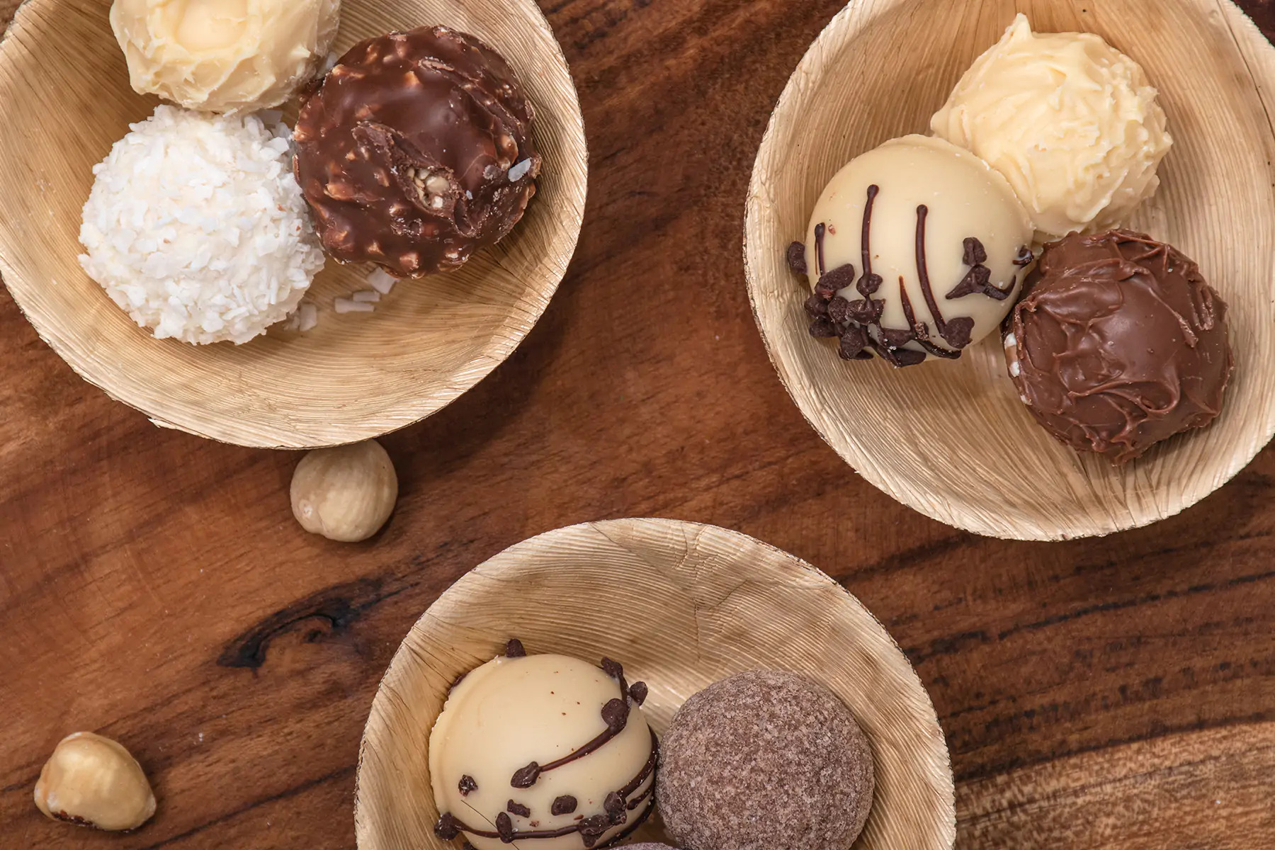 Trzy małe, okrągłe, deserowe kulki o różnej fakturze i kolorze w naturalnych, biodegradowalnych miseczkach z liści palmowych na ciemnobrązowym, drewnianym stole. Dwie orzechy laskowe leżą obok...
