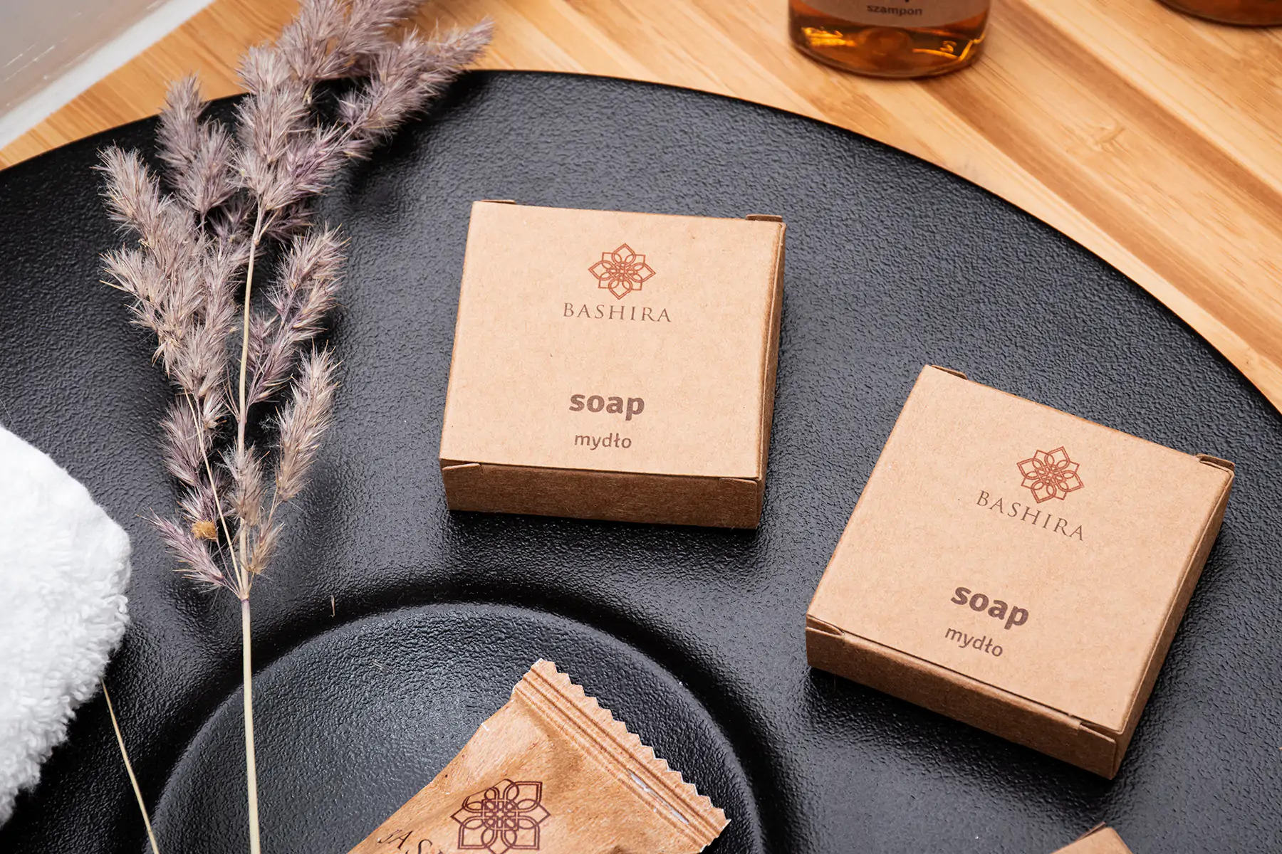 Dwa pudełka mydła 'Bashira' z logo kwiatowym na czarnym, teksturowanym talerzu z dekoracyjną gałązką i fragmentem ręcznika, w tle bambusowa deska.