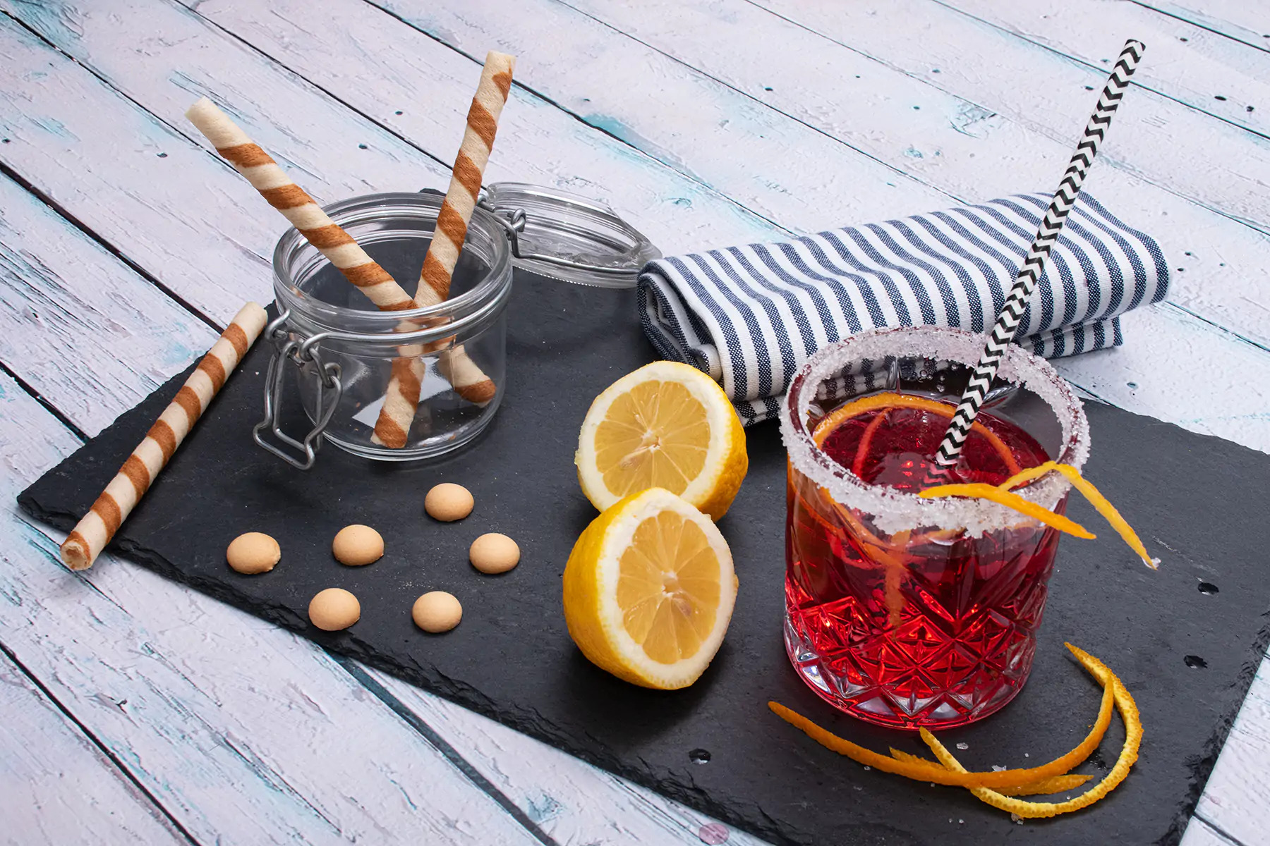 Kompozycja na czarnym kamieniu: drink z krwawych pomarańczy w szklance z cukrem na obrzeżu, obok połówki cytryny, słoik z rurkami waflowymi i ciasteczka na tle drewnianego stołu.