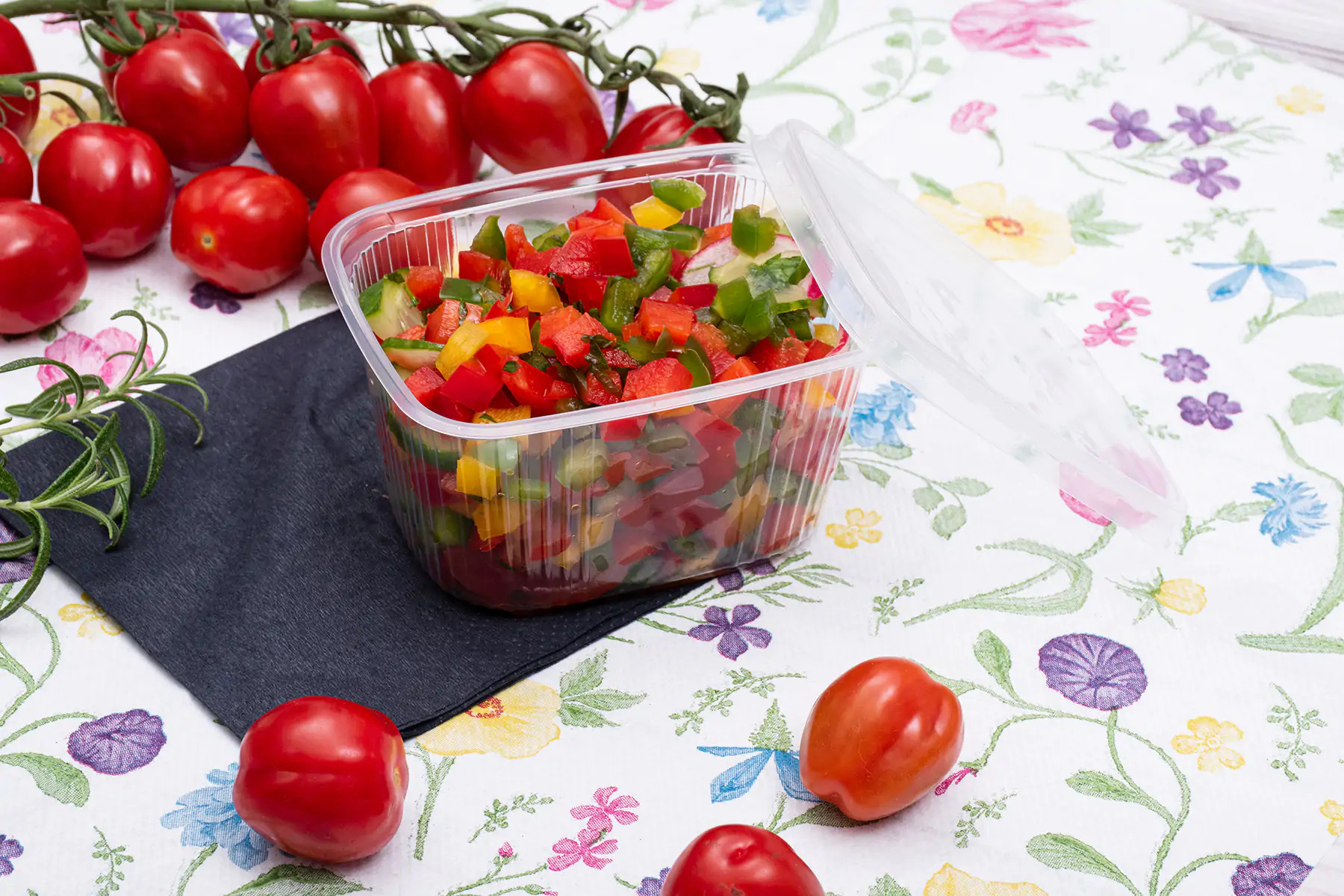 Plastikowe pudełko z pokrojonymi warzywami (papryka, ogórek) na kwiecistej serwetce, obok gałązka pomidorów cherry i czarna serwetka.