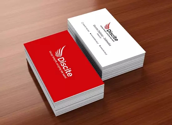 Dwa stosy wizytówek na drewnianym biurku. Jeden stos jest z wizytówkami w kolorze czerwonym z logo firmy, a drugi stos jest z wizytówkami w kolorze białym z danymi kontaktowymi firmy.