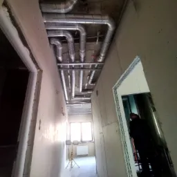 Wnętrze w trakcie prac wykończeniowych: widoczne metalowe rury wentylacyjne pod sufitem, surowe ściany, oświetlenie robocze i fragment sylwetki osoby w przejściu.