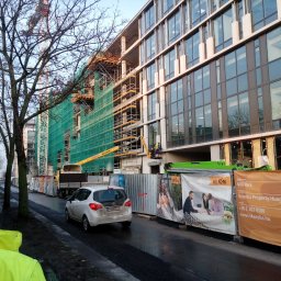 Alu - Prestiż - Budowa budynku biurowego z widocznymi oknami, rusztowaniami i dźwigiem, zabezpieczona siatką ochronną, z pracownikami na podnośniku koszowym oraz banerami reklamowymi w tle.