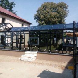 Alu - Prestiż - Nowoczesna konstrukcja z czarnych profili i szkła, prawdopodobnie weranda lub oranżeria, w trakcie budowy, z widocznymi elementami konstrukcyjnymi i stertą płyt betonowych na piasku.