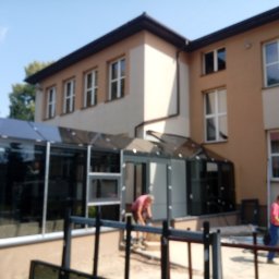 Alu - Prestiż - Montaż przeszklonego wejścia do budynku z oknami, dwóch pracowników w trakcie pracy, widok zza ogrodzenia.