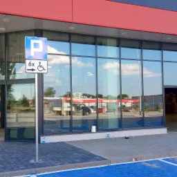 Szklana fasada budynku z automatycznymi drzwiami przesuwnymi i otwartymi drzwiami bocznymi, widoczny znak parkingu dla niepełnosprawnych, odbicie nieba w szybach.