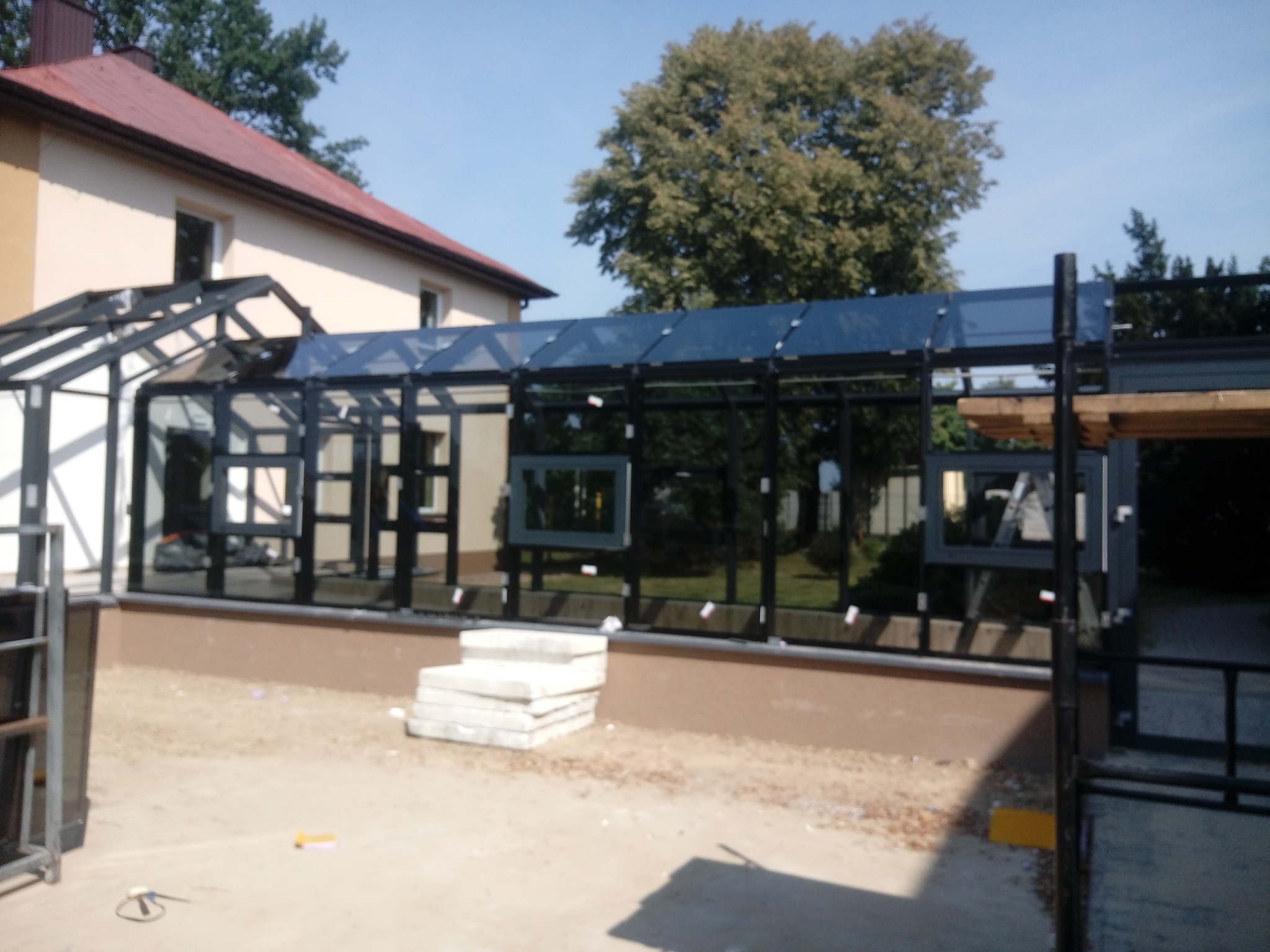 Nowoczesna konstrukcja z czarnych profili i szkła, prawdopodobnie weranda lub oranżeria, w trakcie budowy, z widocznymi elementami konstrukcyjnymi i stertą płyt betonowych na piasku.