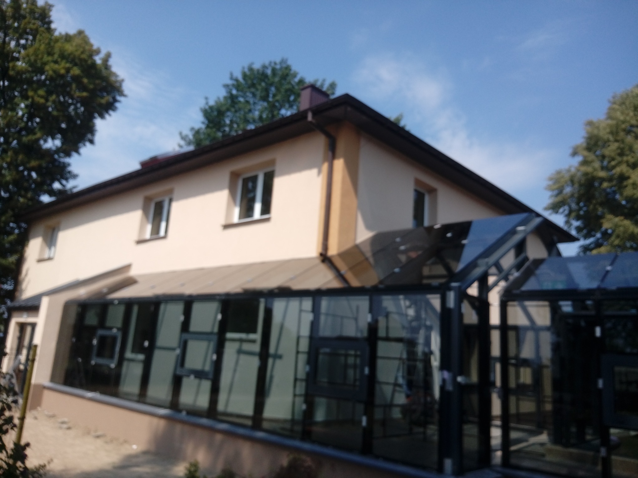 Dwukondygnacyjny dom z dobudowaną przeszkloną oranżerią, widok z zewnątrz w słoneczny dzień.