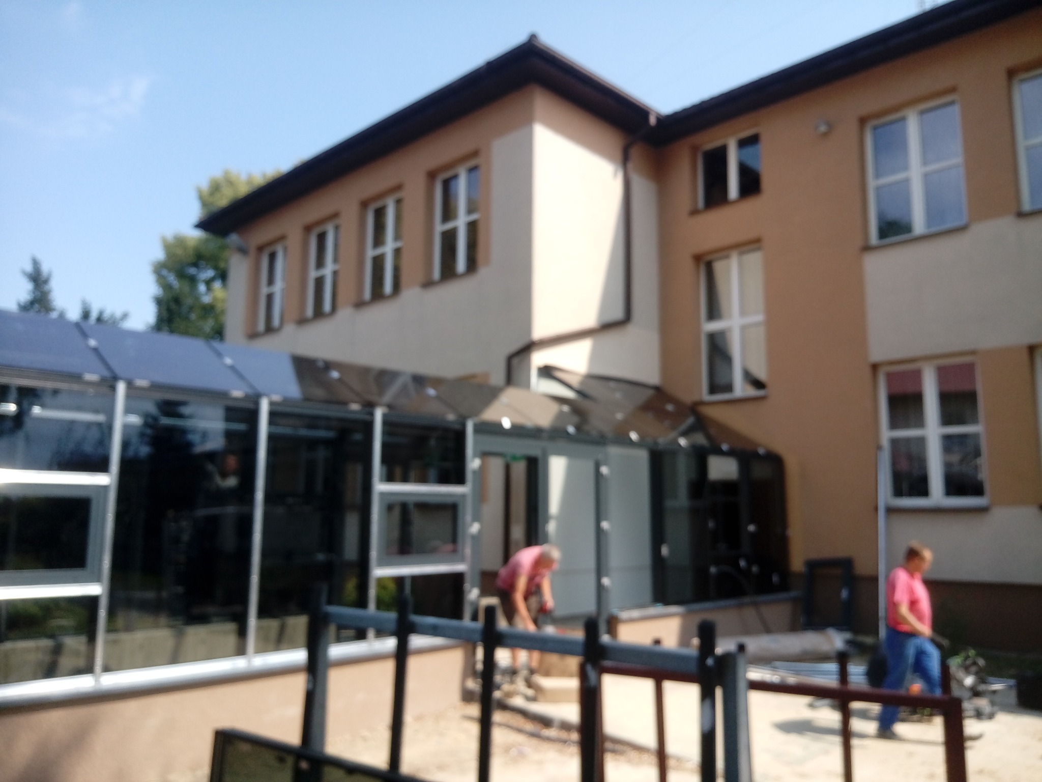 Montaż przeszklonego wejścia do budynku z oknami, dwóch pracowników w trakcie pracy, widok zza ogrodzenia.