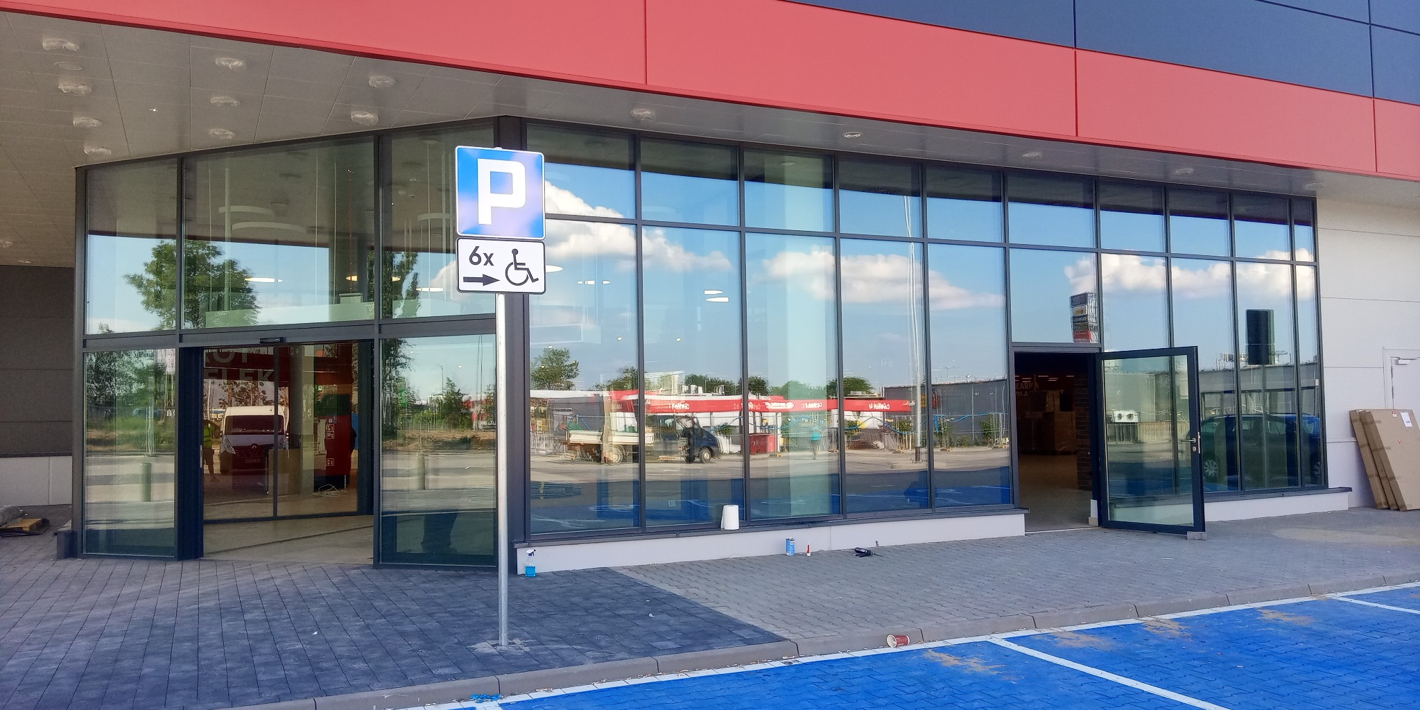 Szklana fasada budynku z automatycznymi drzwiami przesuwnymi i otwartymi drzwiami bocznymi, widoczny znak parkingu dla niepełnosprawnych, odbicie nieba w szybach.